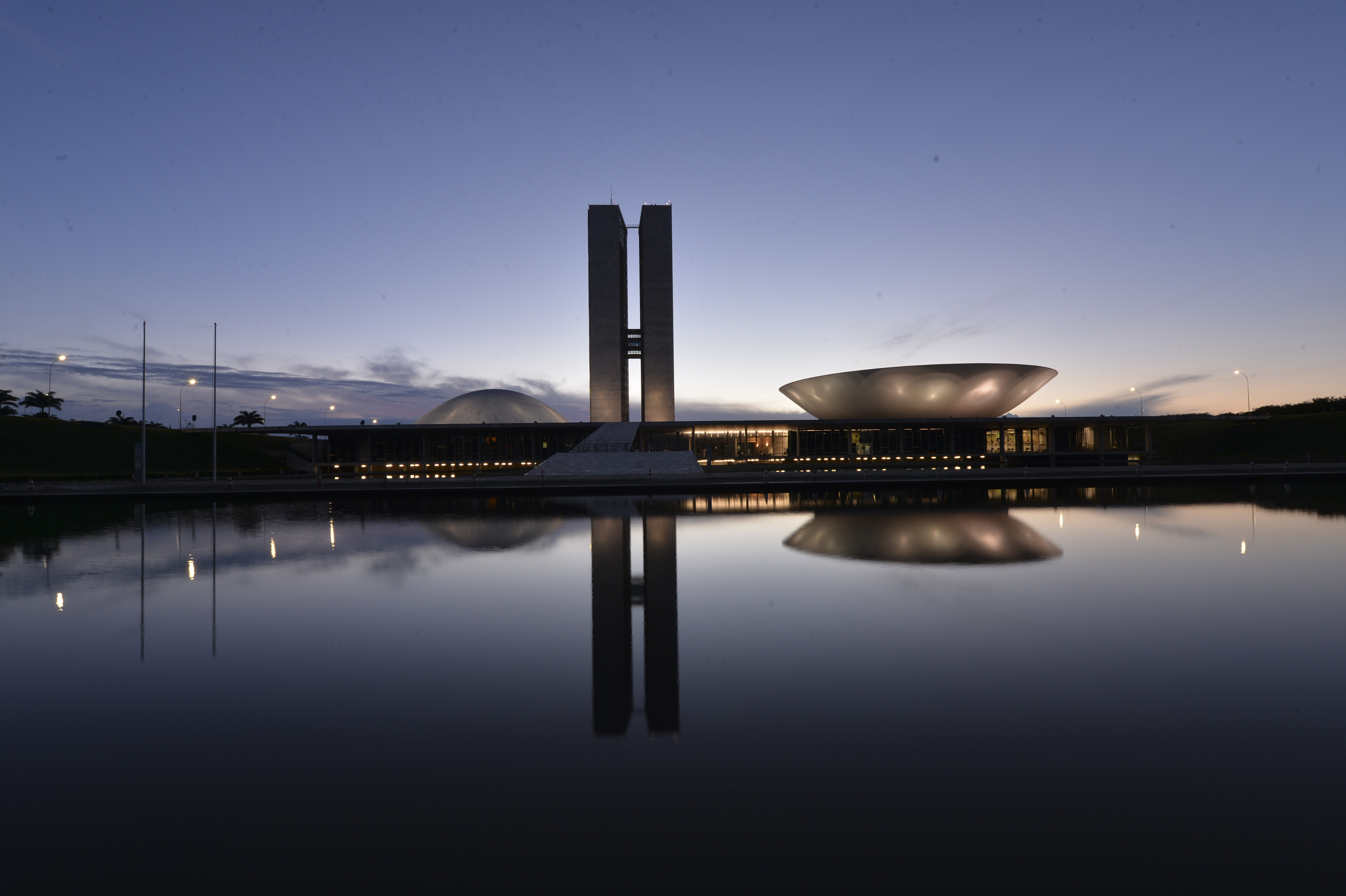Fachada do Congresso Nacional ao entardecer, com as duas torres e os plenários da Câmara e do Senado refletidos no espelho d'água