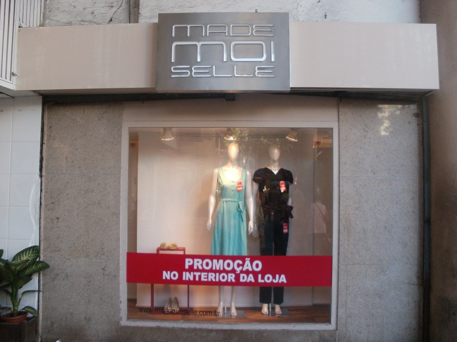 Vitrine de uma pequena loja de roupas, com manequins e um cartaz de promoção, em calçada no Rio de Janeiro