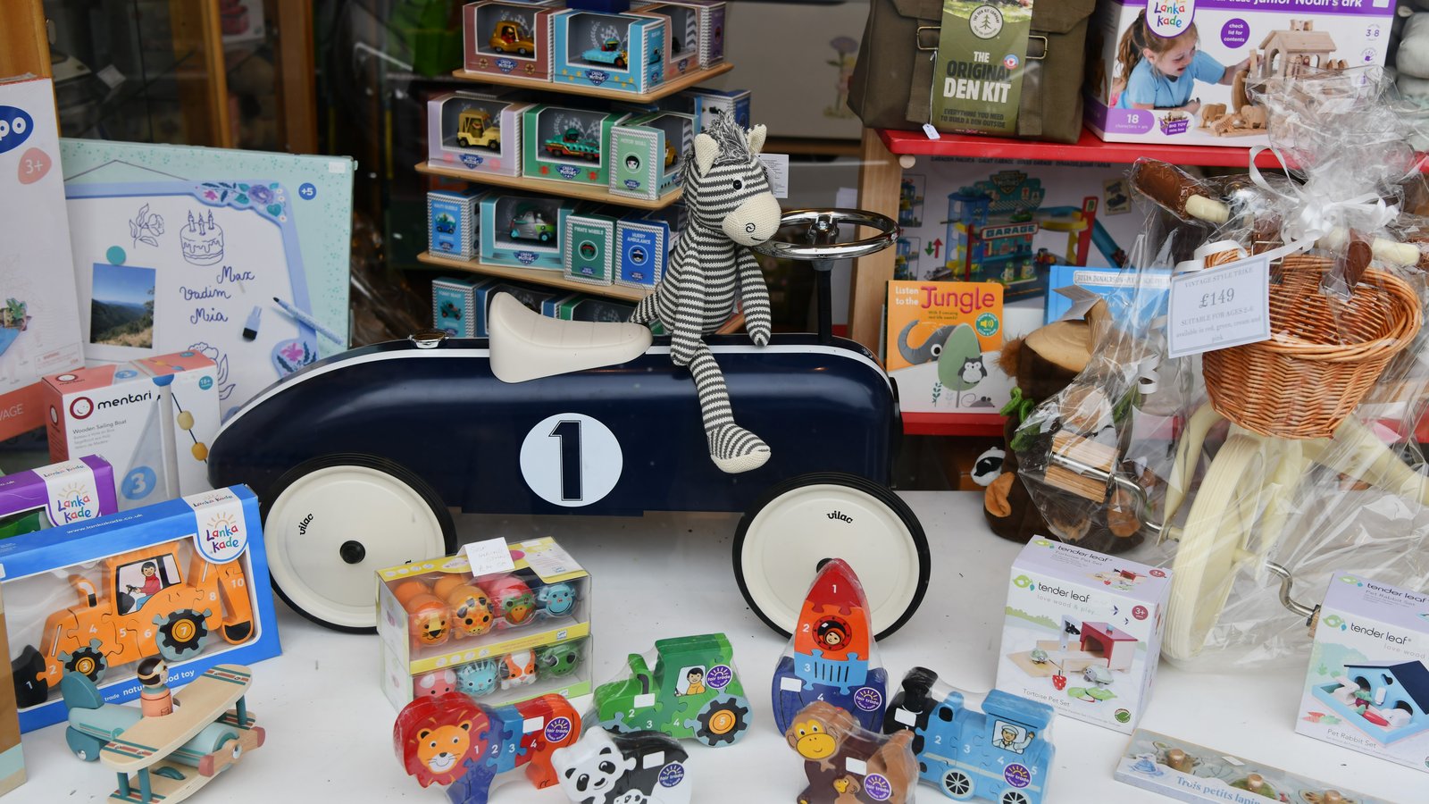 Vitrine de loja de brinquedos com carrinhos, bonecos e brinquedos de madeira expostos, com etiqueta de preço visível